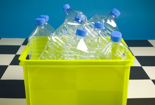 A close-up view of a bright yellow plastic bin filled with clear plastic bottles that have blue screw caps. The bottles are empty, with some partially crumpled, and are placed in a slightly tilted manner within the bin. The background features a black and white checkered surface, possibly a tiled floor or tabletop, contrasting with the vibrant yellow of the bin. The scene appears to be set in an indoor environment, emphasizing the types of waste that a commercial waste removal service like Commercial Waste Chingford would handle, particularly recyclable plastic bottles within a rubbish collection context relevant to Chingford and the surrounding postcode area.