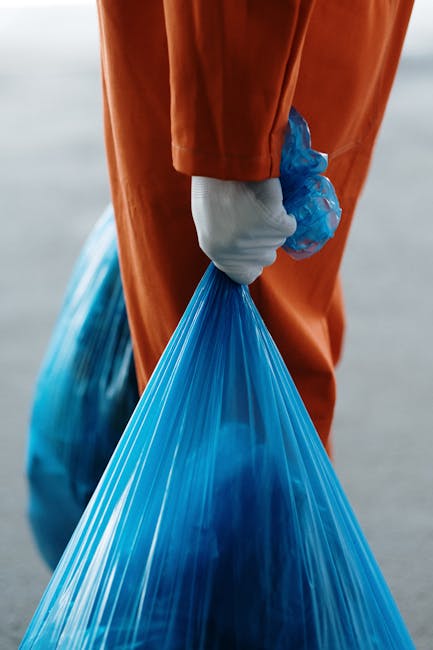 A waste management worker wearing a high-visibility yellow vest and teal pants is operating a street-cleaning or rubbish collection vehicle on an urban street during dusk or early evening. The worker 