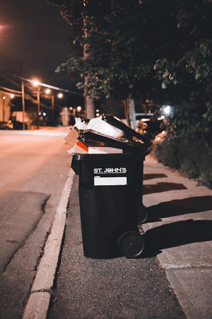A black wheeled rubbish bin labeled 'ST. JOHN'S' positioned on a residential street at night, partially filled with assorted waste including paper and plastic items, some of which are overflowing and 