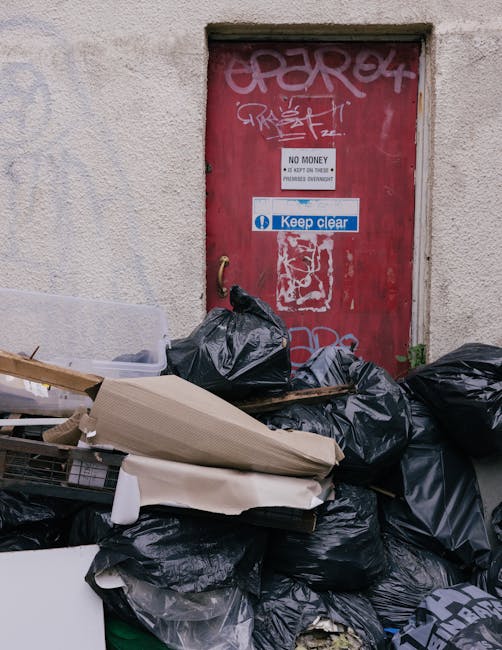 A black wheeled rubbish bin labeled 'ST. JOHN'S' positioned on a residential street at night, partially filled with assorted waste including paper and plastic items, some of which are overflowing and 